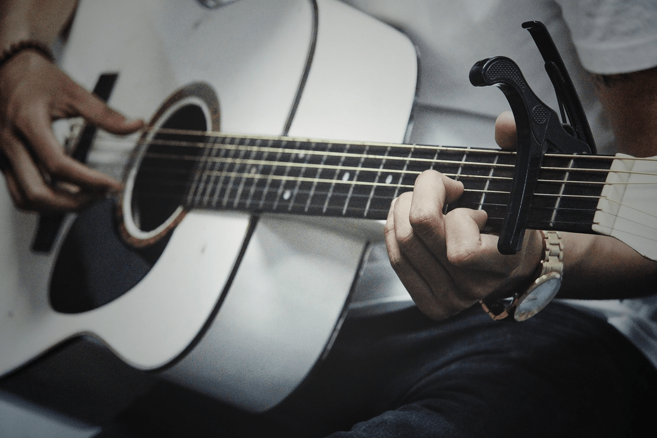 A close up of a man playing a white acoustic guitar using fingerstyle and a capo